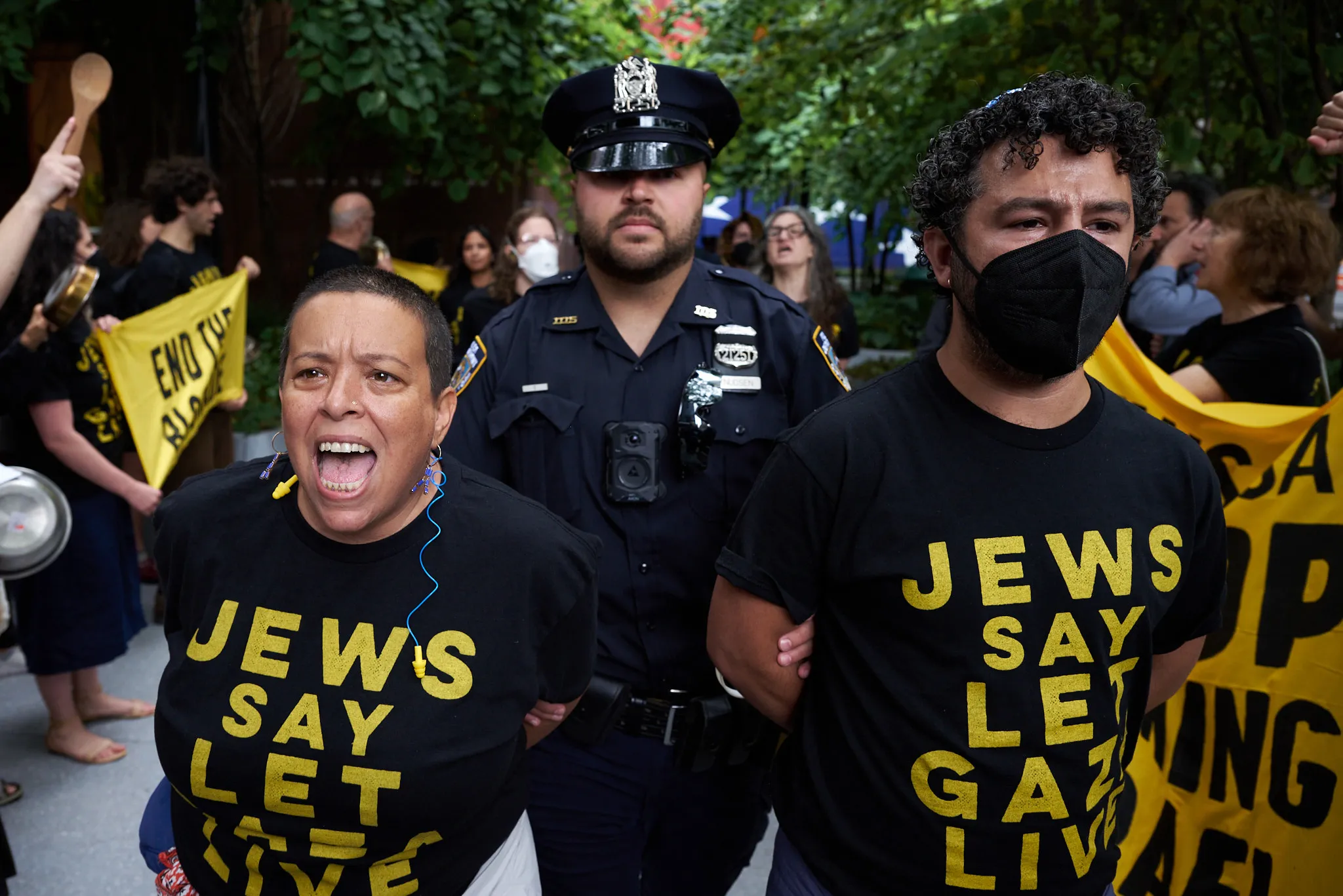 Two handcuffed protestors, one wearing a kippah, wearing shirts that read "Jews say let Gaza live" being led by an NYPD officer