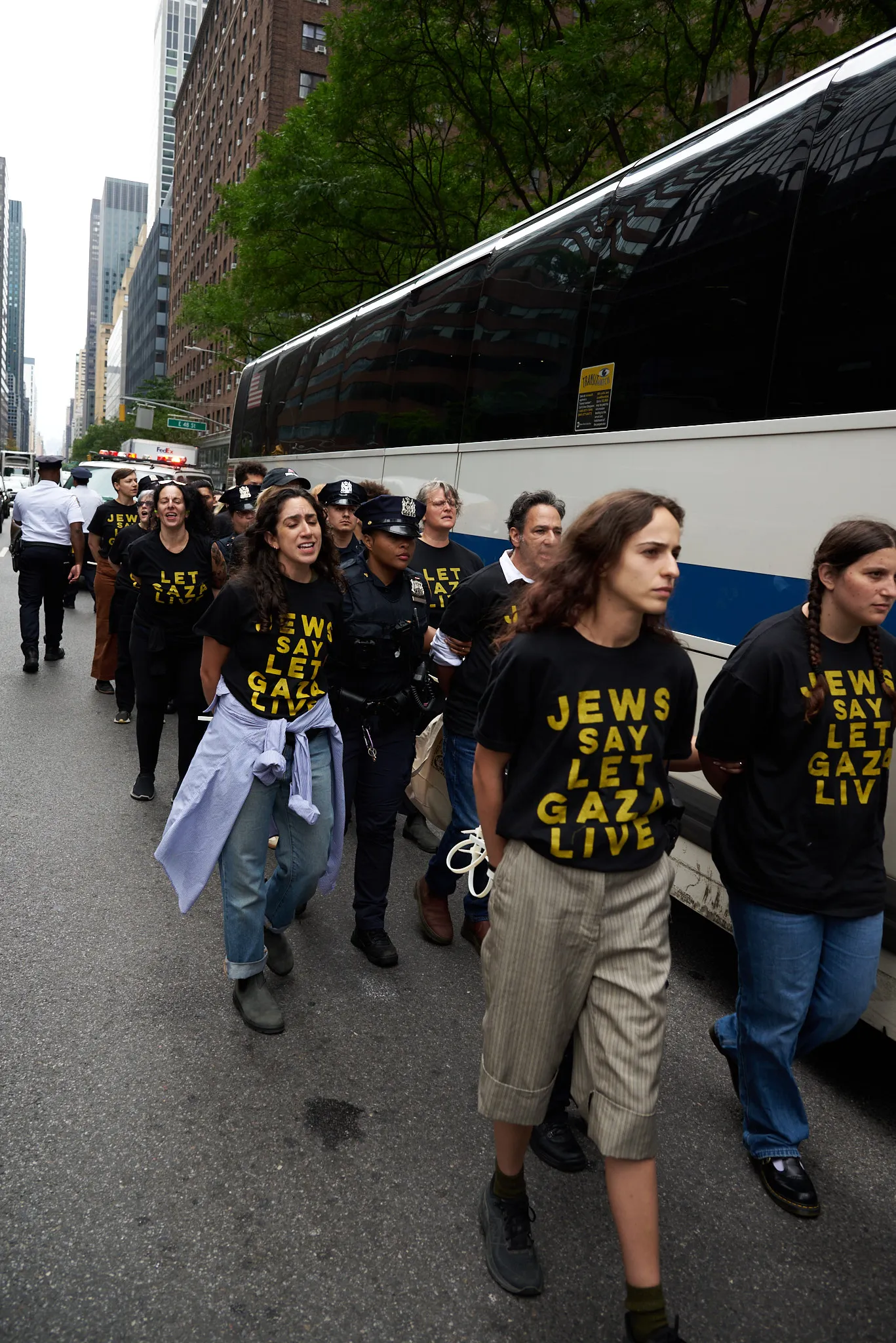 Long line of handcuffed protestors, mainly women, being led towards an MTA bus for transport